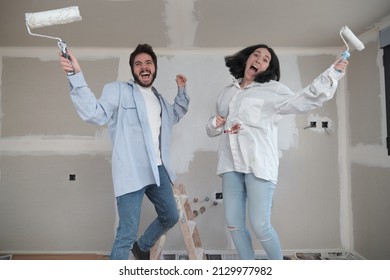 Young Happy Caucasian Couple Jumping With Paint Rollers And Laughing While Painting Walls In Their New House In Construction.