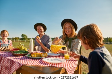 Young Happy Caucasian Hipster Farm Family Having Lunch With Fresh Organic Products In Countryside Outdoors. Mother Pour Juice For Son. Father And Daughter Eat Fruits. Modern Farm Lifestyle. Sunny Day