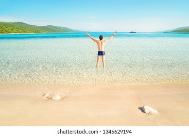 young happy caucasian man wearing speedos standing on beautiful empty Sakarun beach with his back to camera on sunny summer day with yacht in distance, Dugi island, Croatia