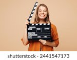 Young happy Caucasian woman she wear orange shirt casual clothes hold in hand classic black film making clapperboard isolated on plain pastel light beige background studio portrait. Lifestyle concept