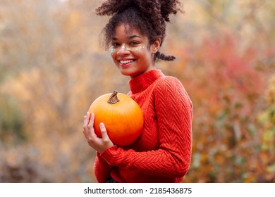 Young Happy Cheerful African American Woman With Curly Hair Holding Orange Pumpkin And Smiling At Camera While Standing Against Blurred Autumn Forest Background. Fall Season And Halloween Concept