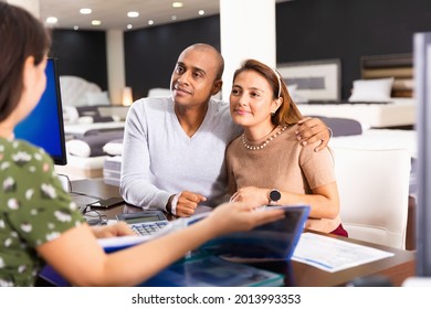 Young happy cheerful hispanic couple consulting with furniture saleswoman when choosing new mattress in store