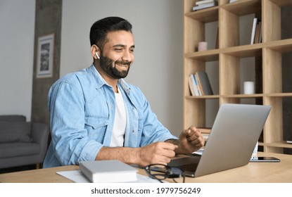 Young happy cheerful indian arab eastern man wearing wireless earphones talking on video conference call using laptop at modern home office. Remote distant online communication technologies concept. - Powered by Shutterstock