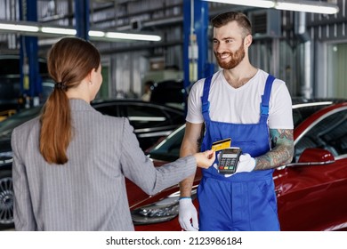 Young Happy Cheerful Professional Car Mechanic Man In Blue Overalls Gloves Hold Payment Terminal Fow Paying With Credit Card By Female Owner Driver Woman Work In Vehicle Repair Shop Workshop Indoors.