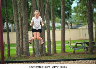 Young Happy Child Girl Playing Alone, Flying High On Swings On Summer Weekend Sunny Day. Safety And Recreation On Playground Concept