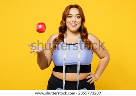 Young happy chubby overweight plus size big fat fit woman wear blue top warm up training hold tape measure eat toss up apple isolated on plain yellow background studio home gym. Workout sport concept
