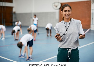 Young happy coach using stopwatch during PE class at school gym and looking at camera. Her students are exercising in the background.