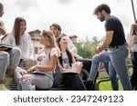 Young, happy, college students having a conversation sitting on a bench in the park out of the campus building, discussing school subjects or project, preparing for exam