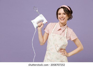 Young happy confident smiling housewife housekeeper chef cook baker woman in pink apron hold mixer stand akimbo arm on waist isolated on pastel violet background studio. Cooking food process concept.