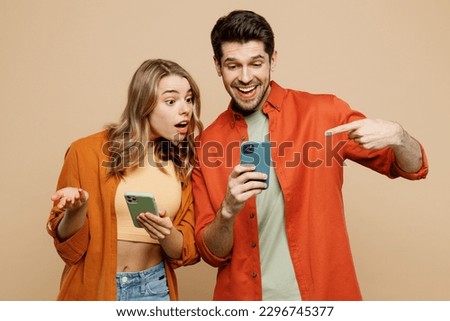 Young happy confused couple two friends family man woman wear casual clothes hold use point finger on mobile cell phone together isolated on pastel plain light beige color background studio portrait