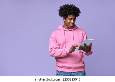 Young happy cool curly African American teenager student boy wearing pink hoodie holding pad using digital tablet computer technology browsing, elearning standing isolated on light purple background.