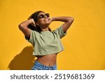 Young happy cool playful African American gen z woman wearing sunglasses having fun, smiling Afro girl standing at yellow color wall background outdoors feeling joy enjoying sunny summer day.