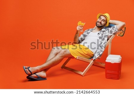 Young happy cool tourist man 20s wear beach shirt hat lie on deckchair near fridge drink exotic cocktail isolated on plain orange background studio portrait. Summer vacation sea rest sun tan concept