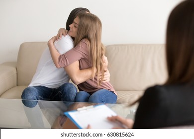 Young happy couple after successful therapy session with family psychologist, sitting on sofa embracing, solving problems in relationships, reconciliation between spouses, mutual understanding