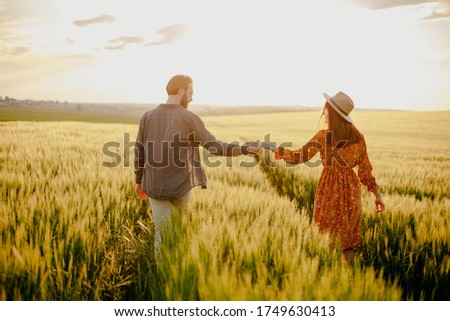 young happy couple in a barley field at sunset. girl in a yellow dress and a guy with a beard. 
portrait of couple walking in the grass