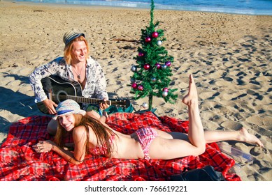 Young happy couple, boy and girl with decorated Christmas tree playing on beach at Christmas time, enjoying the sun and xmas holiday, attractive couple in love celebrating Christmas on the beach party