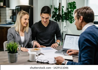 Young happy couple communicating with real estate agent while examining blueprints on a meeting. 