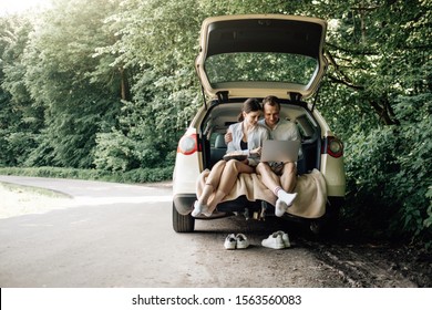 Young Happy Couple Dressed Alike In White T-shirt Sitting In The Car Trunk With Laptop And Popcorn On The Roadside, Weekend Outside The City, Holidays And Road Trip Concept