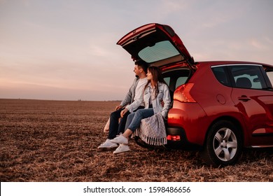 Young Happy Couple Dressed Alike in White Shirt and Jeans Sitting at Their New Car Trunk, Beautiful Sunset on the Field, Vacation and Travel Concept