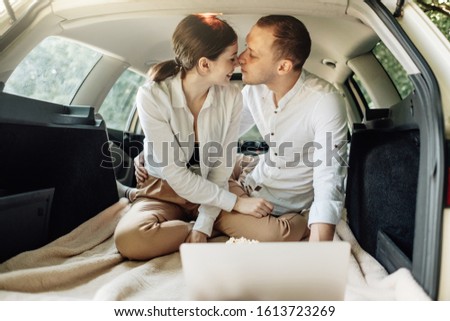 Young Happy Couple Dressed Alike in White T-shirt Sitting in the Car Trunk with Laptop and Popcorn on the Roadside, Weekend Outside the City, Holidays and Road Trip Concept