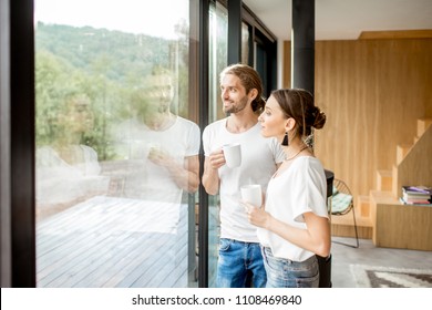 Young and happy couple dressed in white shirts standing together with cups near the window at home