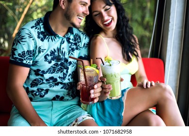 Young Happy Couple Drinking Tasty Sweet Cocktails At Tropical Bar, Smiling And Having Fun, Bright Clothes And Positive Emotions.