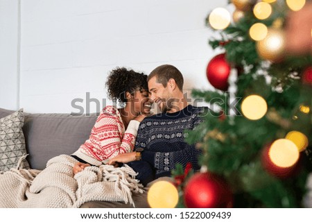 Young happy couple embracing and relaxing on comfortable couch. Lovely multiethnic couple sitting on sofa wearing christmas sweater and hugging. African woman and affectionate man near christmas tree.