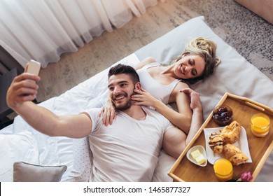 Young happy couple having breakfast in luxury hotel room using smart phone to take selfie photo.