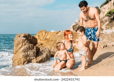 Young happy couple having fun with little kids while enjoying vacation on seashore near waving ocean against rocky mountains - Powered by Shutterstock