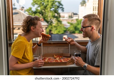 A young happy couple is having meal, dinner, enjoying pizza, salad and drinking Kvass at home on the balcony with open windows, amazing view and portable compact table