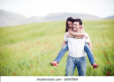 Young Happy Couple With Her At Piggiback Un Spring Field