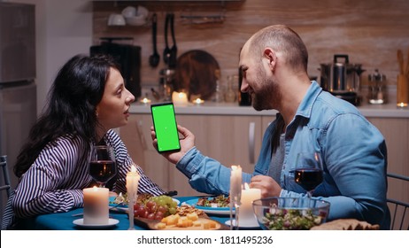 Young Happy Couple Holding Green Screen Phone At Dinner. Cheerful People Looking At Mockup Template Chroma Key Isolated Smart Phone Display Using Techology Internet Sitting At The Table In Kitchen.