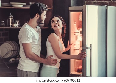 Young Happy Couple Hugging And Smiling While Standing In The Kitchen At Home And Look At The Open Refrigerator.