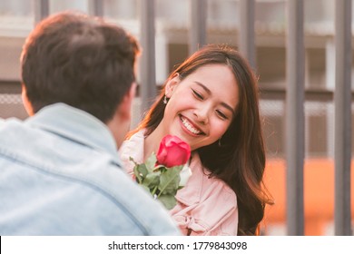 Young Happy Couple Love And Romantic At First Date Relationship. Asian Teenage Woman Surprise And Smiling At Boyfriend Gives Red Rose Flowers At Dinner In Valentine Day. Couple And Happiness Concept.