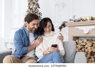 Young happy couple man and woman sitting at home on sofa hugging near Christmas tree and using mobile phone, shopping online, watching video. - Powered by Shutterstock