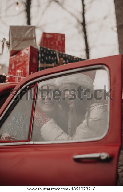 Young happy couple man and woman in love in\
Christmas hats hugging and sitting in a red retro car with New\
Year\'s gifts. Kiss, girl, happiness, quarantine Christmas\
celebration, holiday,\
coronavirus.