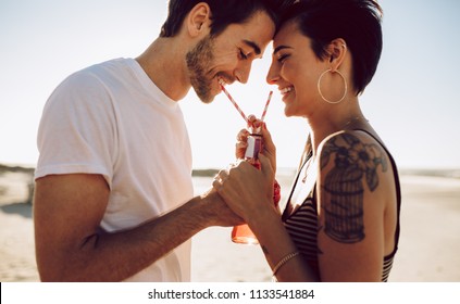 Young Happy Couple Outdoors Drinking Cold Drink From Glass Bottle With Straw. Side View Of Man And Woman Drinking Soft Drink And Smiling.