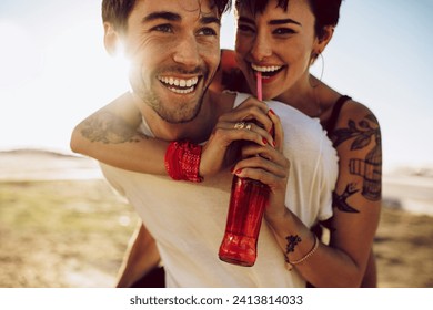 Young happy couple outdoors having fun. Man carrying his girlfriend on his back drinking soft drink from glass bottle with straw. - Powered by Shutterstock