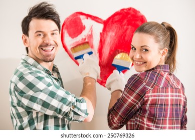 Young Happy Couple Are Painting A Heart On The Wall While Doing Repair At Home.