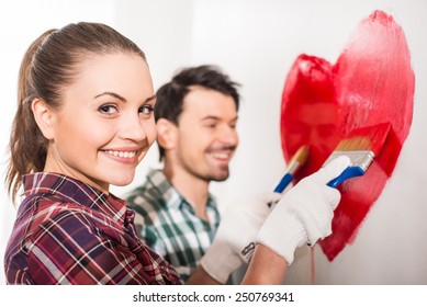 Young Happy Couple Are Painting A Heart On The Wall While Doing Repair At Home.