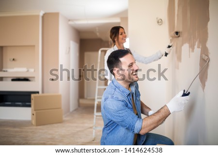Young happy couple painting walls with painting roller in light brown color while wearing gloves.
