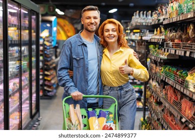 Young happy couple posing in supermarket with shopping trolley cart, lady showing thumbs up, spouses embracing and smiling at camera - Powered by Shutterstock