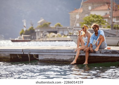 A young happy couple is relaxing with the drink while sitting on the dock on the seaside with their legs in the water on a beautiful sunny day. Love, relationship, holiday, sea