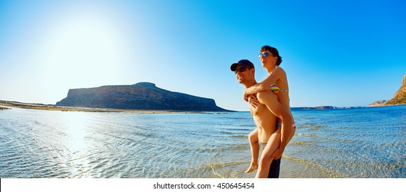 Young Happy Couple Running On The Beach, Woman Sitting On A Man's Back. Balos Beach, Crete, Greece.