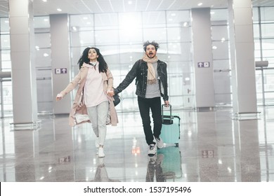 Young Happy Couple Running Through White Empty Airtport. Late On Flight. Worried About Not Being On Time. Vacation Ususal Trip. Unhappy And Scared. Holding Hands Together. Suitcase Behind