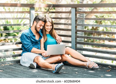 Young Happy Couple Shopping Online On Their Laptop Sitting On Wooden Floor Outside - Two Lovers Watching A Video On A Notebook Computer And Surfing On Internet  - Love, Technology And People Concept