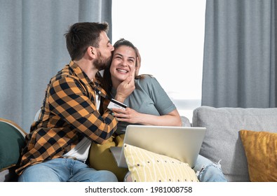 Young Happy Couple Sitting At Home Man Rejoices Because His Wife Made Him Happy With A Gift She Bought Online Via Laptop Computer And Paid With A Credit Card For Their Wedding Anniversary