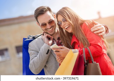 Young Happy Couple With Smart Phone And Shopping Bags In The City