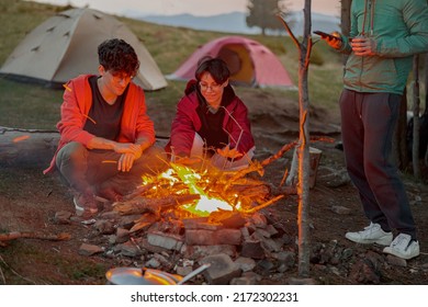 Young Happy Couple Of Travelers Sitting At Fire In Evening At Camping Place. Romantic Moment.