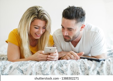 Young Happy Couple Using Their Smartphones. Man And Woman With  Mobile Phone In The Morning Lying On The Bed At Home. Millenial Relationship With Technology Concept.